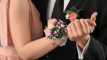 Момиче в прасковена рокля с гривна от розови и бели цветя на китката държи ръката на момче в черен костюм с бутониера.